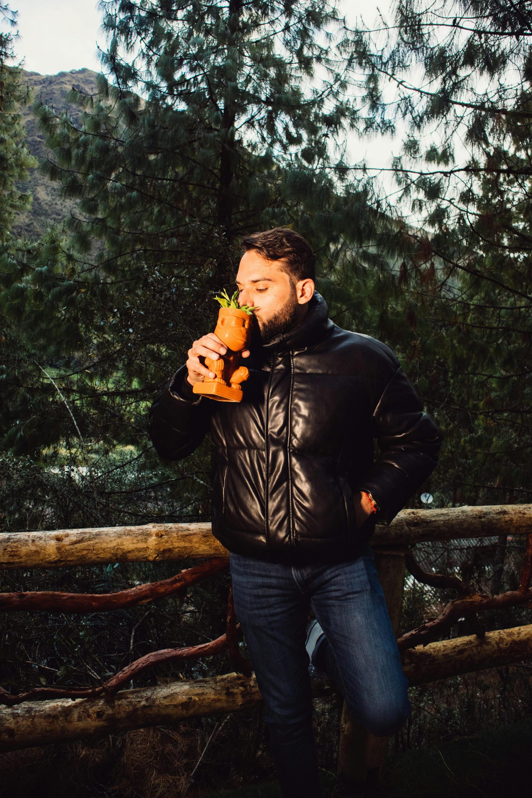 Un homme en veste noire savoure une boisson dans une forêt, entouré d'arbres imposants.
