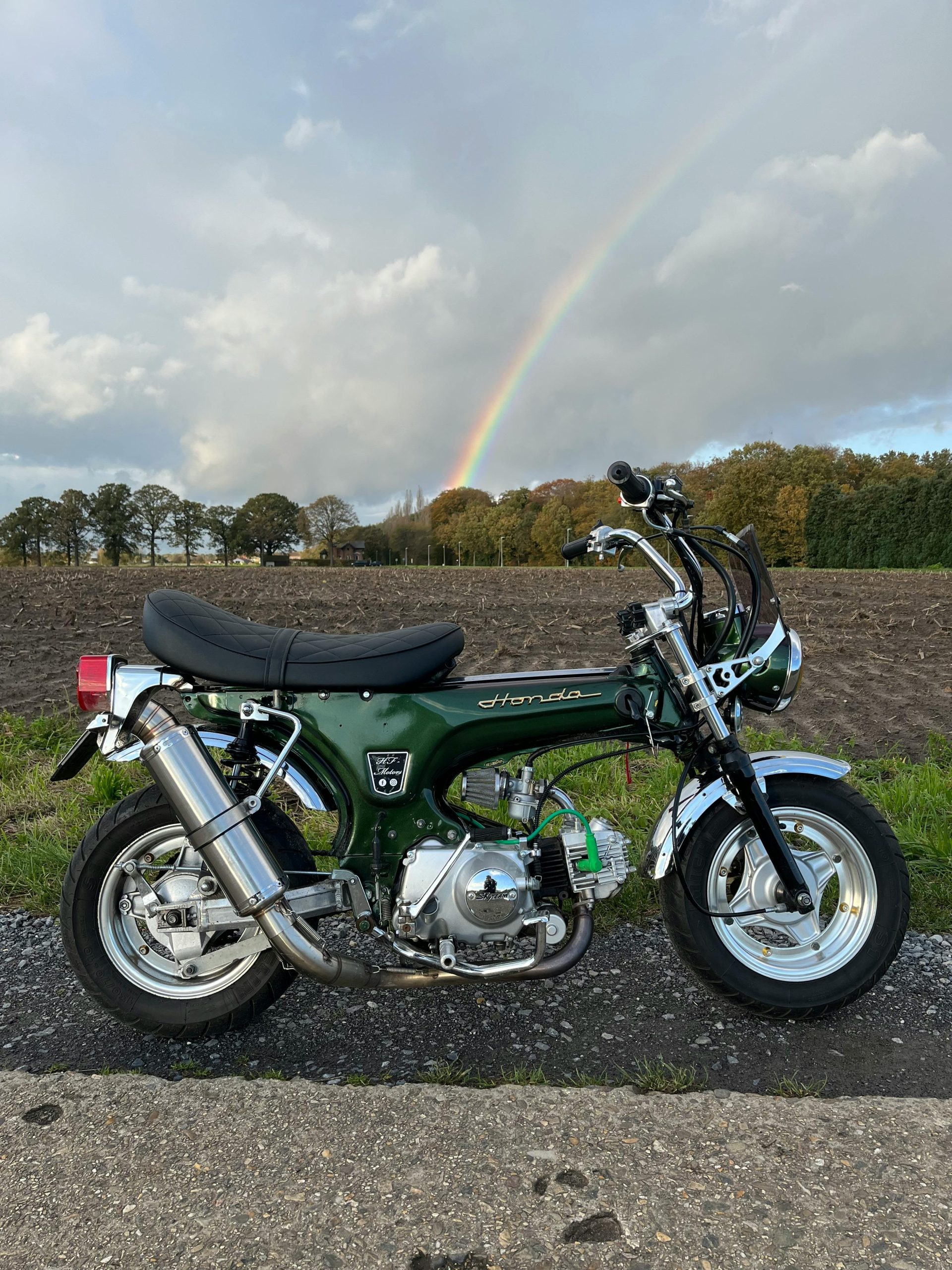 Green Honda Dax motorbike sur une route rurale avec un arc-en-ciel en arrière-plan pittoresque.