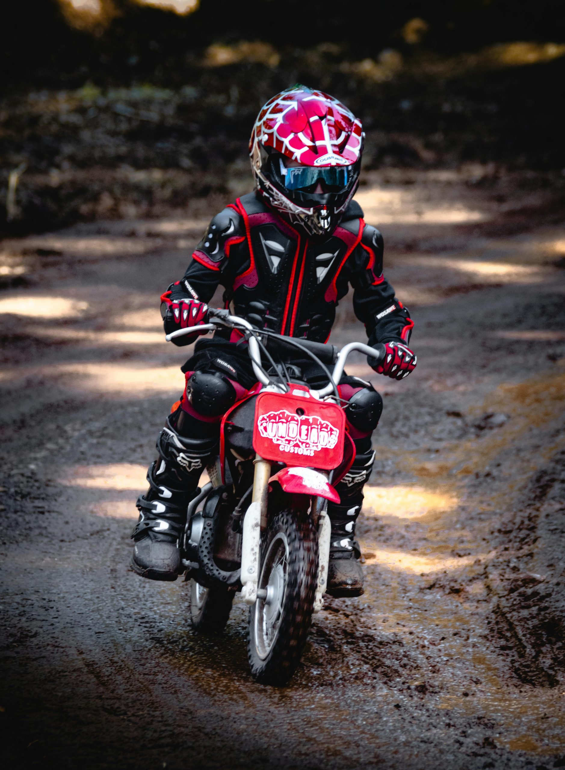 Jeune enfant en équipement de protection roulant sur une mini moto cross sur un sentier boisé.