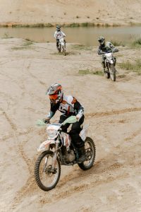 Three motocross riders navigating sandy terrain by a lake, showcasing off-road skill and adventure. Trois motocyclistes de motocross naviguant sur un terrain sablonneux près d'un lac, démontrant leur habileté hors route et l'aventure.