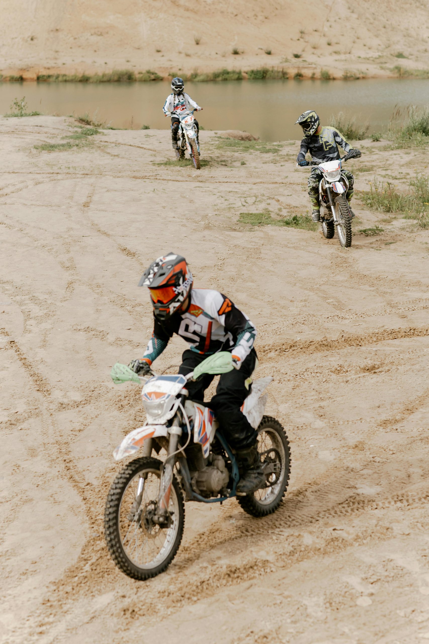 Three motocross riders navigating sandy terrain by a lake, showcasing off-road skill and adventure. Trois motocyclistes de motocross naviguant sur un terrain sablonneux près d'un lac, mettant en avant les compétences tout-terrain et l'aventure.