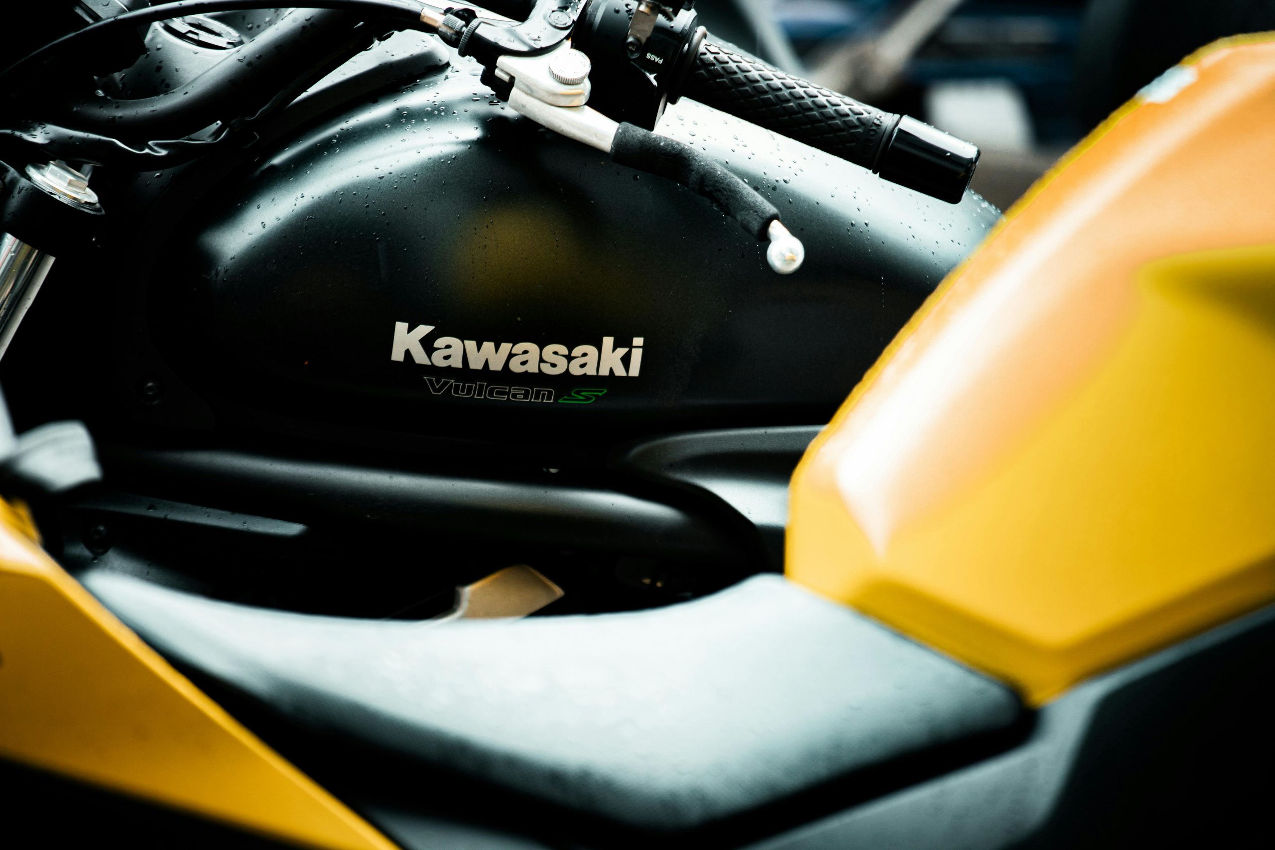 Close-up of a Kawasaki Vulcan S motorcycle's tank and handlebar with raindrops, highlighting its sleek design. Gros plan sur le réservoir et le guidon d'une motocyclette Kawasaki Vulcan S avec des gouttes de pluie, mettant en évidence son design élégant.