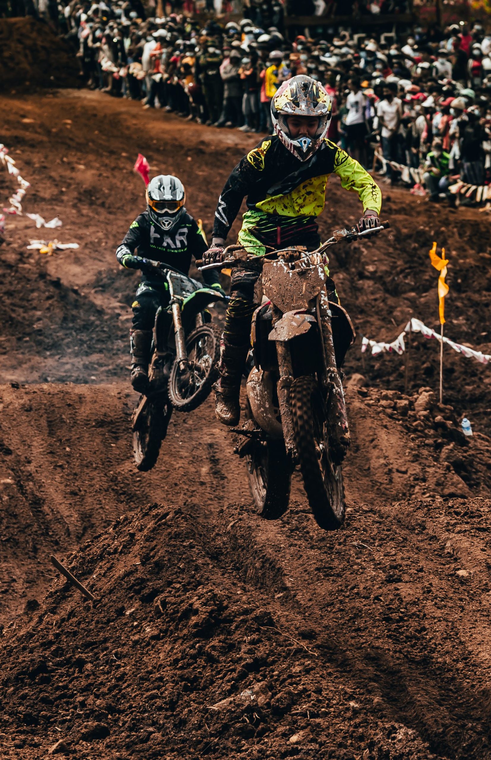 Two motocross riders racing on muddy track with spectators. Deux motocyclistes motocross se disputant une course sur une piste boueuse avec des spectateurs.