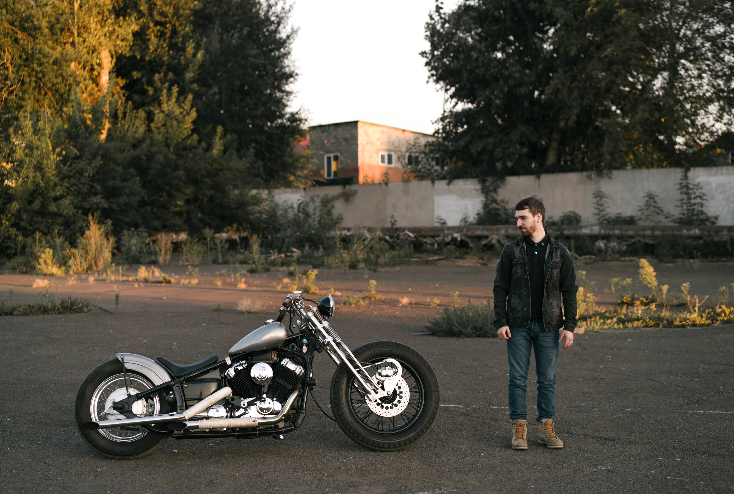 A man stands beside a custom vintage motorcycle in an outdoor setting during the day. Un homme se tient à côté d'une moto vintage personnalisée dans un cadre extérieur pendant la journée.
