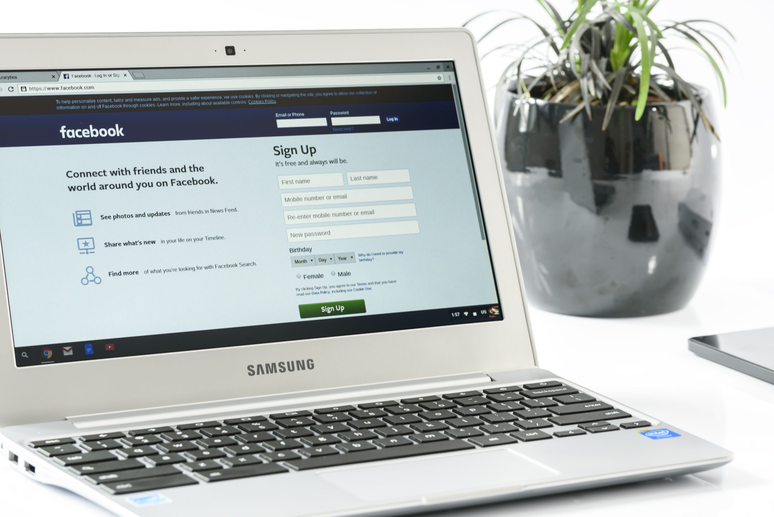 Open Samsung laptop showing Facebook sign-up page next to a potted plant. Ideal for technology themes. Un ordinateur portable Samsung ouvert montrant la page d'inscription Facebook à côté d'une plante en pot. Idéal pour les thèmes technologiques.