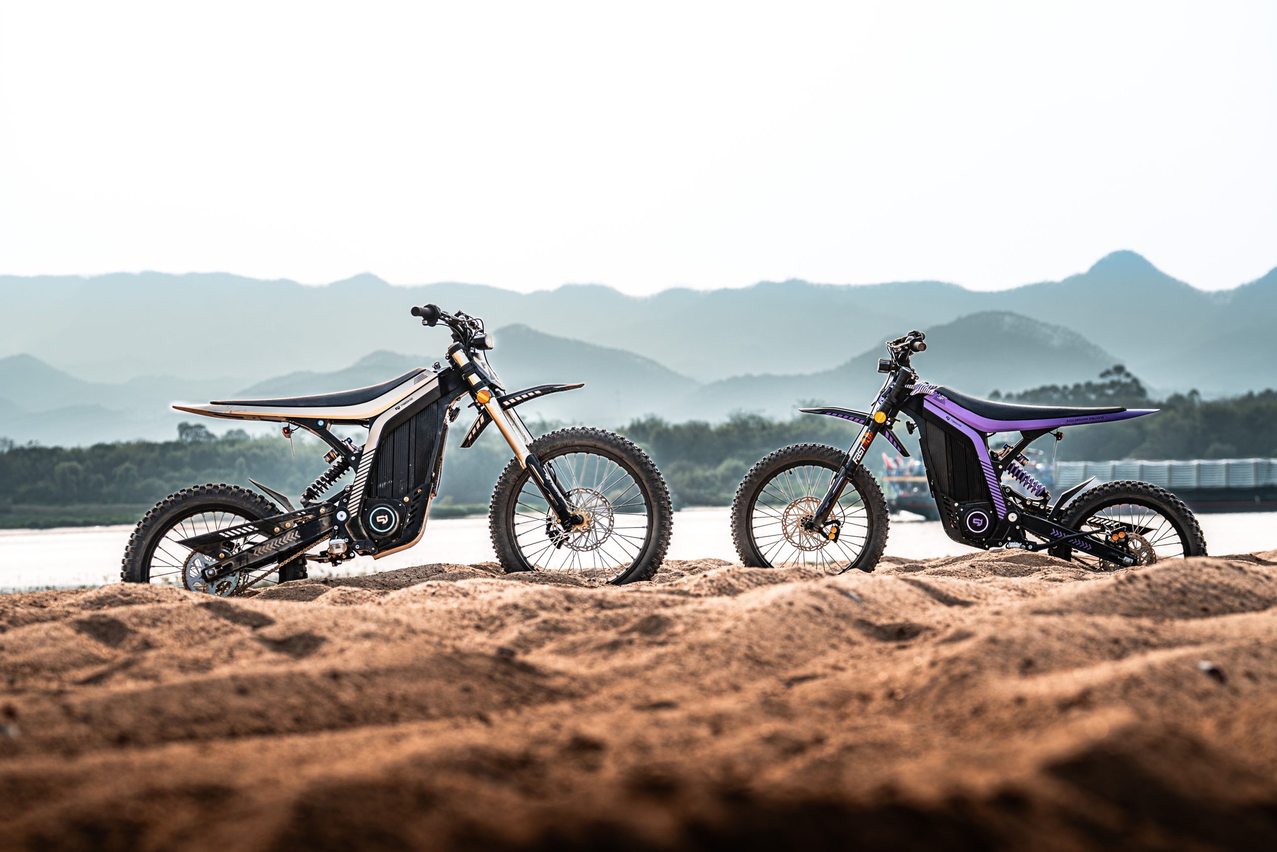 Two electric dirt bikes parked on sandy terrain against a scenic mountain backdrop during sunrise. Deux vélos électriques tout-terrain garés sur un terrain sablonneux avec un paysage montagneux spectaculaire en arrière-plan au lever du soleil.