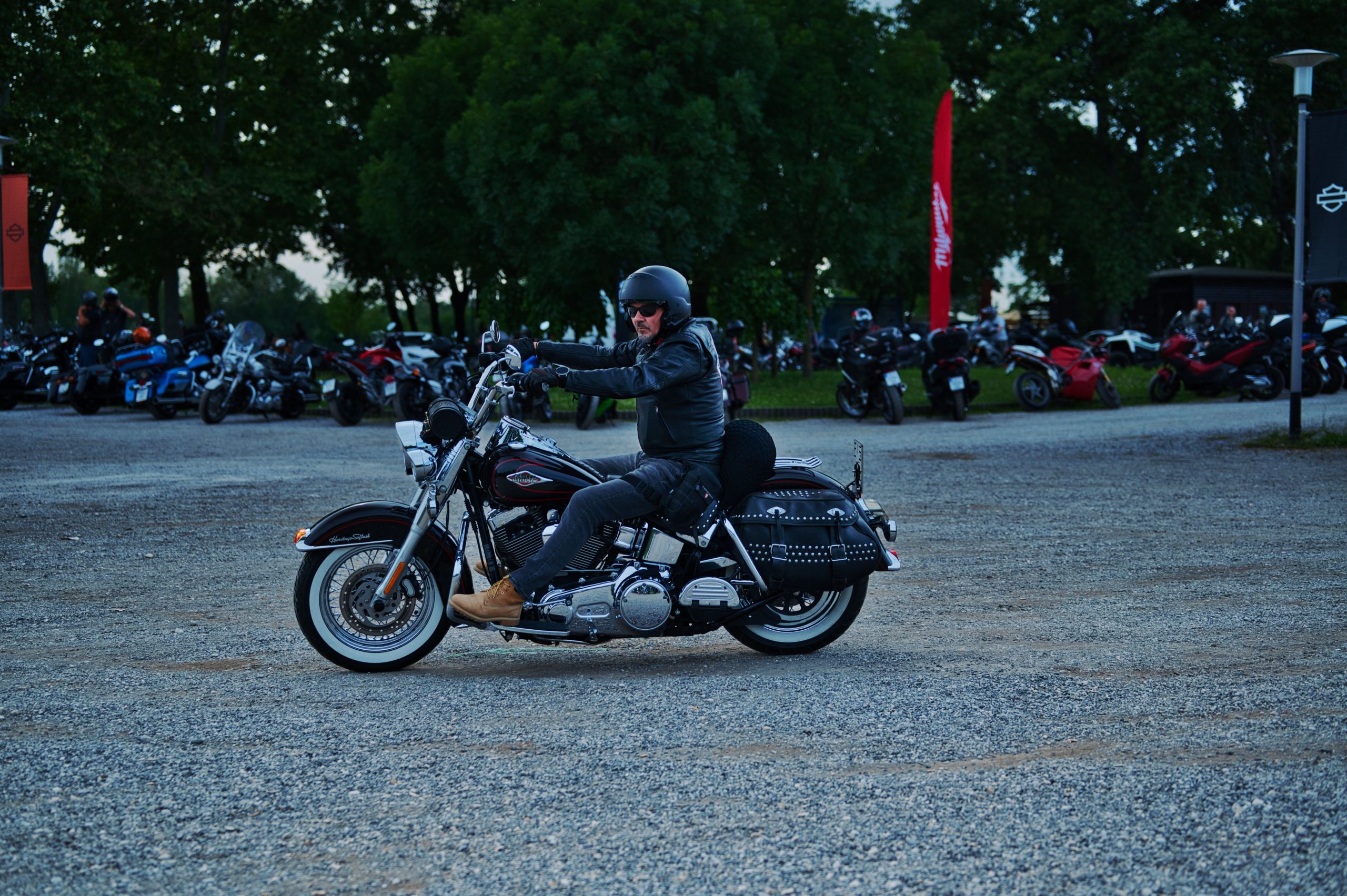 A biker riding a Harley-Davidson at a motorcycle gathering outdoors, surrounded by trees. Un motard chevauchant une Harley-Davidson lors d'un rassemblement de motocyclistes en plein air, entouré d'arbres.