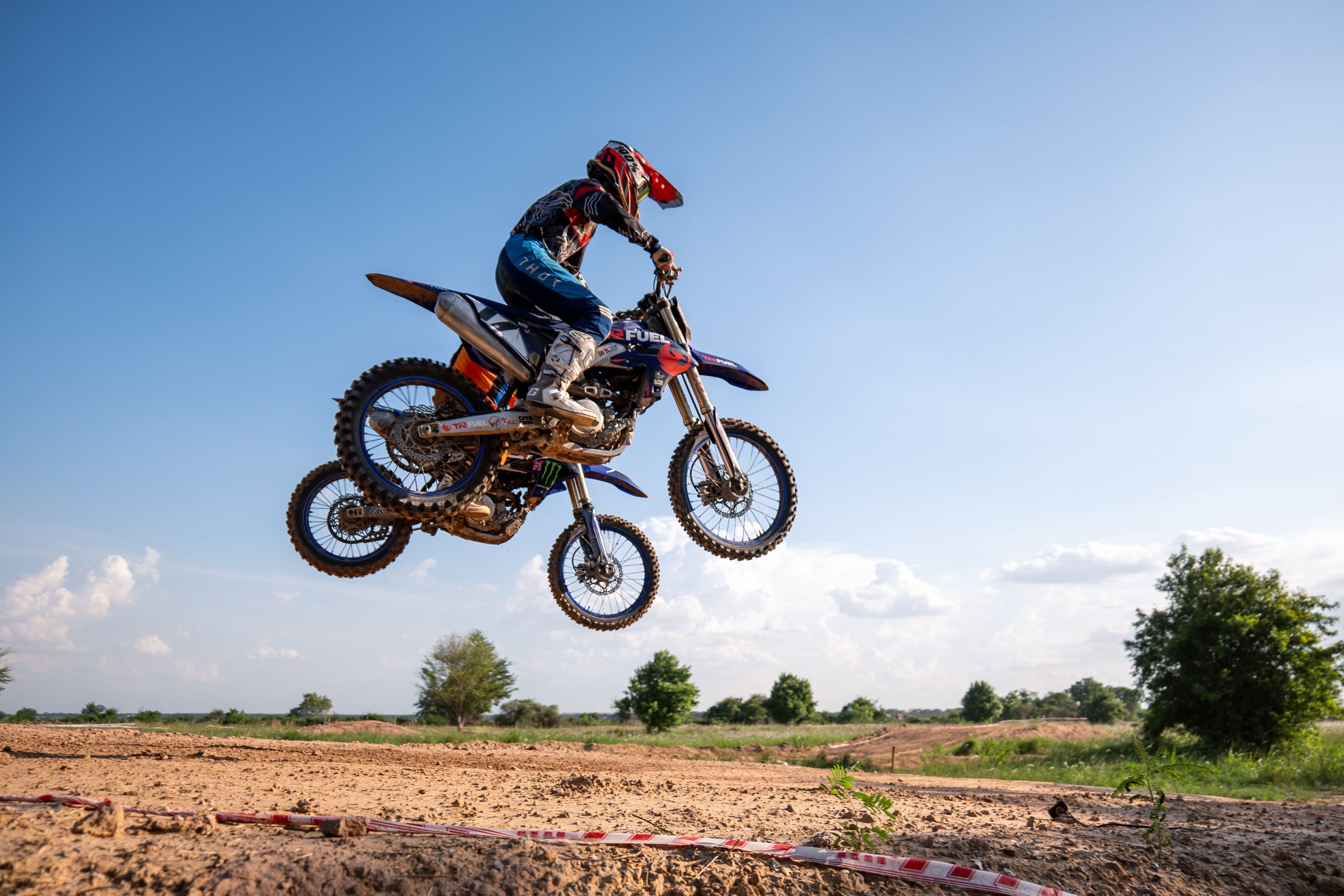 Motocross rider performs a thrilling jump on an outdoor dirt track under clear skies. Le pilote de motocross effectue un saut palpitant sur une piste de terre extérieure sous un ciel dégagé.