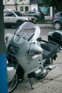 Close-up of a sleek silver motorcycle parked outdoors, featuring distinct design elements and urban background. Gros plan d'une élégante moto argentée garée à l'extérieur, présentant des éléments de design distincts et un arrière-plan urbain.